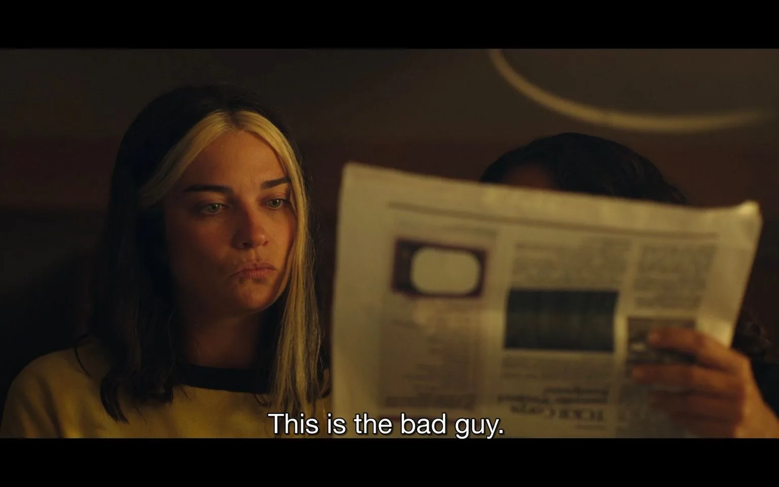 The image features a scene set indoors with dim lighting, creating an intimate and somewhat tense atmosphere. In the foreground, a person with distinctive hair—dark with prominent light streaks—sits with a focused yet pensive expression. They are wearing a yellow top, and their gaze is fixed intently on a newspaper, which is held up by another person partially obscured from view. The newspaper is in the center of the frame, suggesting it contains significant information pertinent to the scene. The caption at the bottom, "This is the bad guy," implies a moment of revelation or identification, possibly in a narrative context such as a film or TV show. The overall color palette is warm, with browns and yellows dominating the scene, enhancing the mood of quiet intensity.