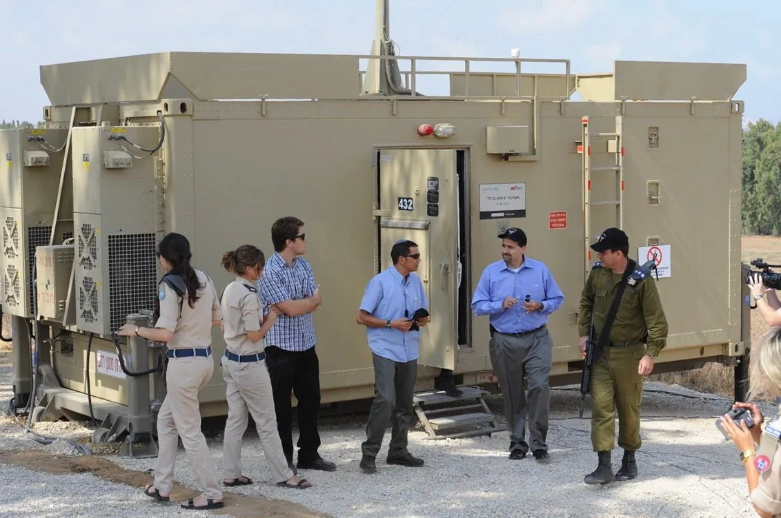 The image shows a group of people standing outside a military-style installation or unit marked with the number "432." The location appears to be outdoors, possibly at a military base or a similar setting. The structure is a large, tan-colored, rectangular building with visible vents and electrical connections, suggesting it's used for operational purposes.
Among the group, there are individuals in military-style uniforms, indicating a formal or official visit. Some are wearing berets, and one person seems to be holding a rifle, emphasizing the military context. There are also civilians present, dressed in casual attire, engaged in conversation. The atmosphere seems professional and slightly informal, with people holding notepads or other small items, possibly for documentation purposes.
In the background, there's a natural landscape with trees, adding to the sense of a remote or controlled area. The lighting is bright, suggesting daytime, and the overall mood appears focused and purposeful.