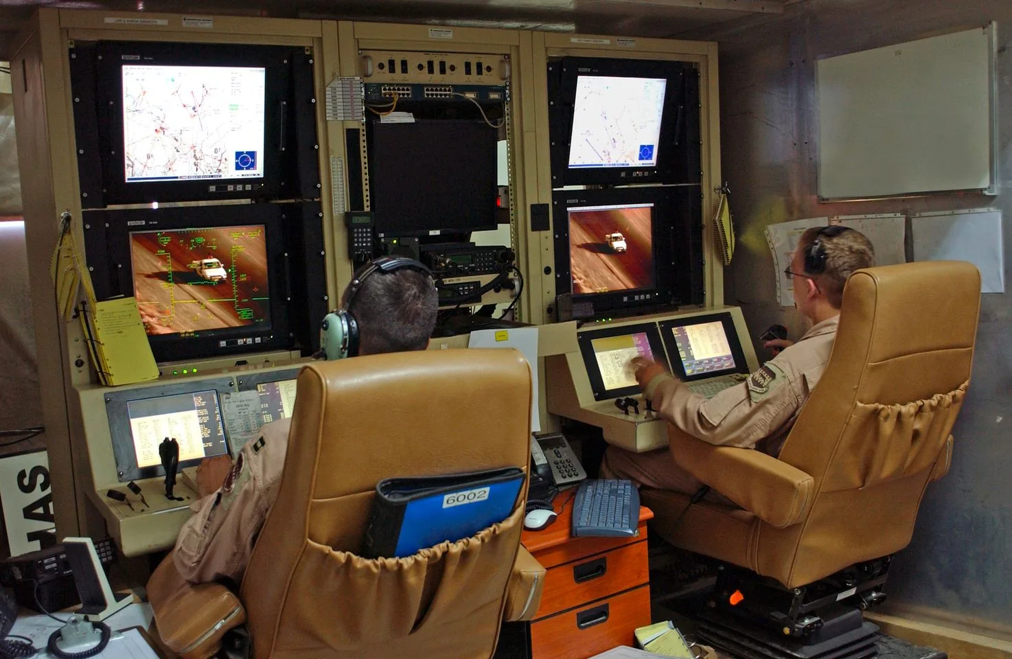 The image depicts a military control room with two operators seated in front of an array of monitors and control panels. The operators are focused on their tasks, wearing headsets and military uniforms, indicating a high-tech and professional environment. The room is filled with advanced equipment, including multiple screens displaying maps, aerial imagery, and other data, suggesting a surveillance or UAV (unmanned aerial vehicle) control station.
The operators are seated in tan, cushioned chairs, indicating comfort for long periods of operation. One of them appears to be interacting with a touchscreen, highlighting the integration of modern technology in their operations. The lighting in the room is subdued, creating an atmosphere of focus and concentration. There is a noticeable presence of communication devices, such as telephones and headsets, indicating constant connectivity and the need for quick information exchange.
A small shelf holds binders and papers, suggesting the need for additional reference materials during operations. The environment is organized, with a mixture of digital and analog tools, reflecting the complexity and precision required in military control scenarios. The presence of maps and real-time imagery on the monitors highlights the critical nature of their work, potentially involving real-time decision-making and coordination.