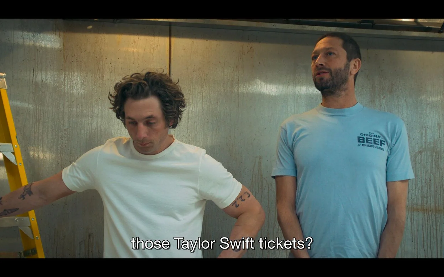 This image is a screenshot from a scene featuring two men, one of whom is identified as Richard 'Richie' Jerimovich, played by Ebon Moss-Bachrach. They are standing in an indoor setting that appears to be a workplace environment. The background consists of a textured wall, suggesting an industrial or kitchen setting.
The man on the left is wearing a plain white t-shirt, and his posture is relaxed, leaning slightly against a yellow ladder. He has curly hair and a contemplative expression, suggesting he is lost in thought or considering something carefully. The man on the right is wearing a light blue t-shirt with text on it that says, "THE ORIGINAL BEEF OF CHICAGOLAND," hinting at a possible connection to a restaurant or a brand. His demeanor appears more engaged in the conversation, as he looks towards the other man with a slight hint of curiosity or inquiry.
The lighting is soft and neutral, creating a casual and realistic ambiance. The subtitle at the bottom of the image reads, "those Taylor Swift tickets?" indicating the content of their conversation, which might involve travel plans, events, or personal interests.