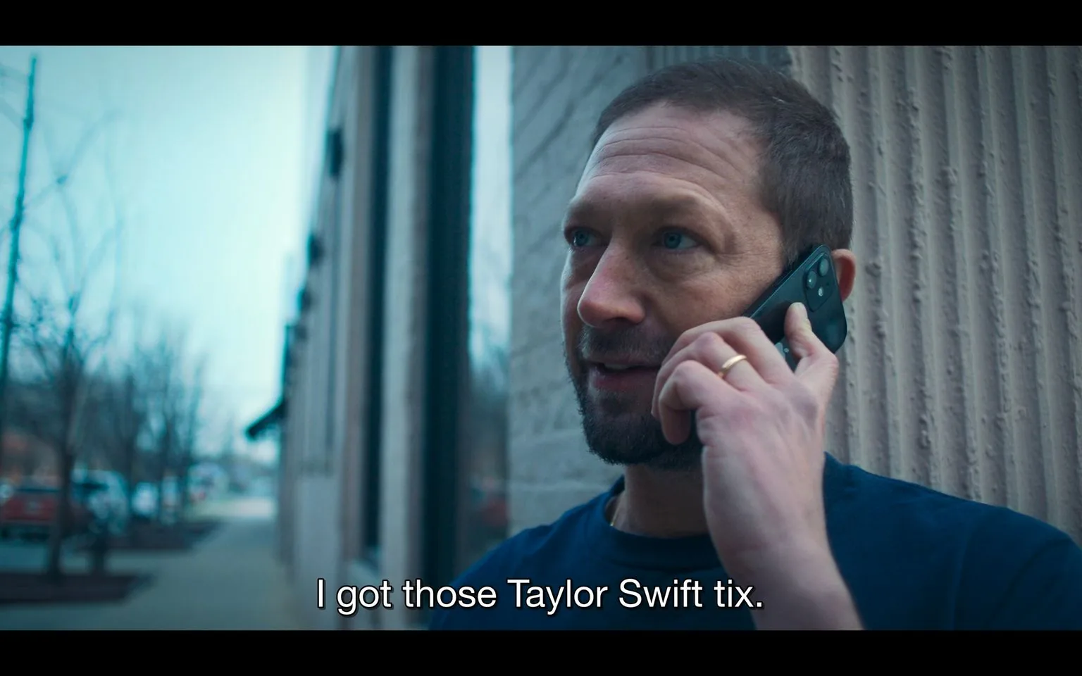The image depicts a man outdoors, standing beside a building with a textured wall. He is engaged in a phone call, holding a smartphone to his ear with his right hand. The setting appears to be a city street, as blurred trees and buildings can be seen in the background. The lighting suggests it is daytime, with a cool blue hue dominating the scene.
The man has a beard, short hair, and is wearing a dark blue shirt. His expression seems focused and slightly pleased, possibly due to the successful acquisition of something desirable. A gold wedding ring is visible on his left hand. The caption at the bottom of the image reads, "I got those Taylor Swift tix," indicating that he has obtained tickets to a Taylor Swift concert, which is likely a moment of excitement or relief. The framing focuses closely on his upper body and face, emphasizing the personal nature of the conversation. The image captures a casual yet significant moment, hinting at the cultural popularity and demand for Taylor Swift concert tickets.