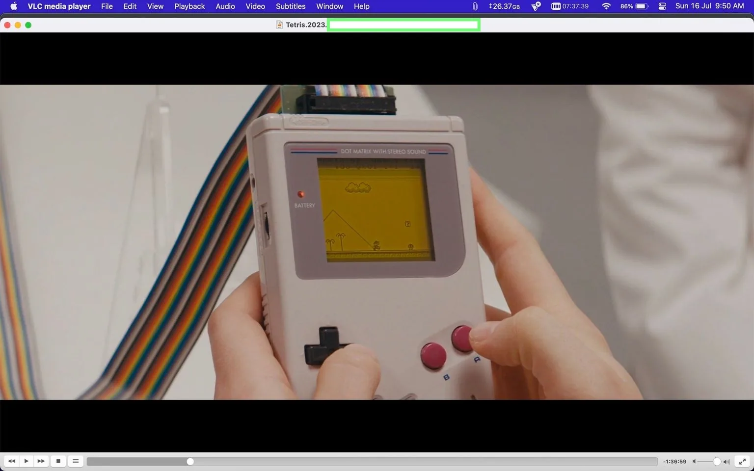 In this screenshot, a scene from a film titled "Tetris.2023" is playing on VLC media player. The focus is on a vintage handheld gaming device, a Game Boy, being held by a pair of hands. The Game Boy features a small screen displaying a game with simple graphics, including elements like clouds, a pyramid, and palm trees. The device has a distinctive gray body with a directional pad on the left and two red buttons labeled "A" and "B" on the right. A multicolored ribbon cable is connected to the top of the device, hinting at some technical modification or testing.
The screen text reads "DOT MATRIX WITH STEREO SOUND" at the top and includes a "BATTERY" indicator on the left side. The scene is lit brightly, emphasizing the details of the Game Boy and its screen. The background is blurred but features a white or light-colored setting, possibly an indoor environment like an office or a tech lab. The overall mood is nostalgic, evoking the classic era of handheld gaming.