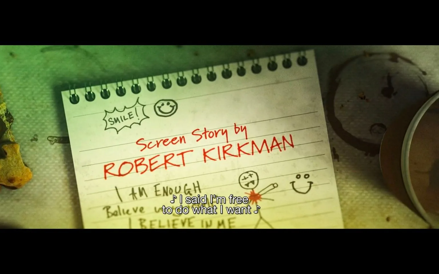 The image is a close-up of a notebook page with a playful and casual aesthetic. The page features the text "Screen Story by ROBERT KIRKMAN" written prominently in red, suggesting a creative or narrative work attributed to him. There are various doodles and motivational phrases like "SMILE!" and "I AM ENOUGH," alongside sketches of smiley faces and a stick figure. The style is informal and handwritten, conveying a relaxed and personal tone.
The setting seems to be a desk or table, with a cup partly visible on the right and a textured surface underneath the notebook. The lighting is slightly dim with a greenish tint, adding a unique mood to the scene. The notebook is spiral-bound, and additional text like "Believe" and "I BELIEVE IN ME" suggests themes of self-affirmation. Subtitles at the bottom of the image say, "♪ I said I'm free to do what I want ♪", indicating that this frame might be from a video or film featuring a musical element.