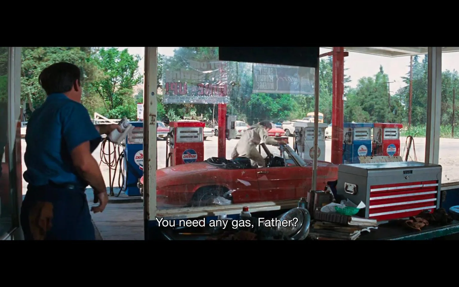 ```
The scene captures a moment at a gas station, with a mechanic dressed in a blue uniform stepping out from the service area. He is holding a white cloth, suggesting he may be ready to assist with a car service. The focus is on a red convertible parked at one of the pumps. A man in a light-colored outfit is getting out of the car. The setting is a typical mid-20th-century gas station, featuring Chevron branding visible on the pumps. The subtitle reads, "You need any gas, Father?" indicating a casual conversation likely directed at the man in the car, hinting he might be a priest due to the term "Father." The scene is set during the daytime with a clear view of greenery and other vehicles in the background, suggesting a rural or suburban environment. The composition includes various details like a metal toolbox and other maintenance equipment in the foreground, enhancing the authenticity of the gas station setting. This screenshot is from the film "The Exorcist," known for its iconic and suspenseful scenes.