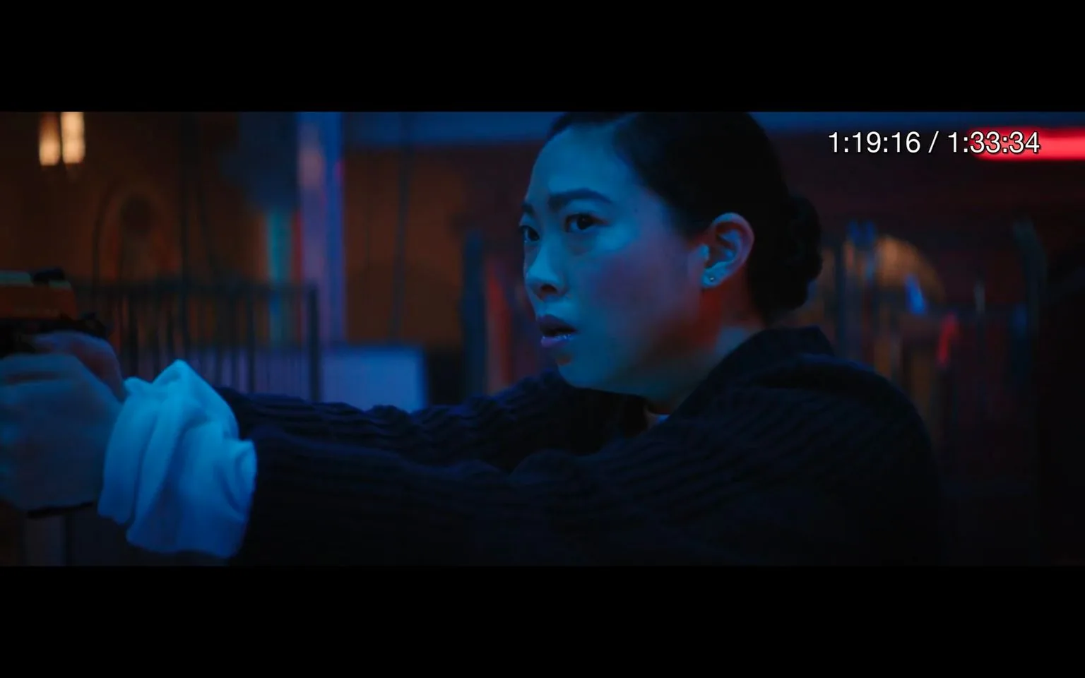 This video frame captures a tense moment indoors with dramatic lighting, primarily in shades of blue and red, creating a moody atmosphere. The focus is on a person holding a gun, aiming intently at something off-screen. The individual is wearing a dark, ribbed sweater with a white undershirt visible at the cuffs, suggesting a casual yet prepared appearance. Their facial expression is one of concentration and determination, indicating they are in a serious or possibly dangerous situation. The setting appears to be dimly lit, with blurred elements in the background, enhancing the sense of focus and urgency in the foreground. The on-screen text shows a timestamp, suggesting this is a scene from a film or series, with the total duration of the video being 1 hour and 33 minutes.