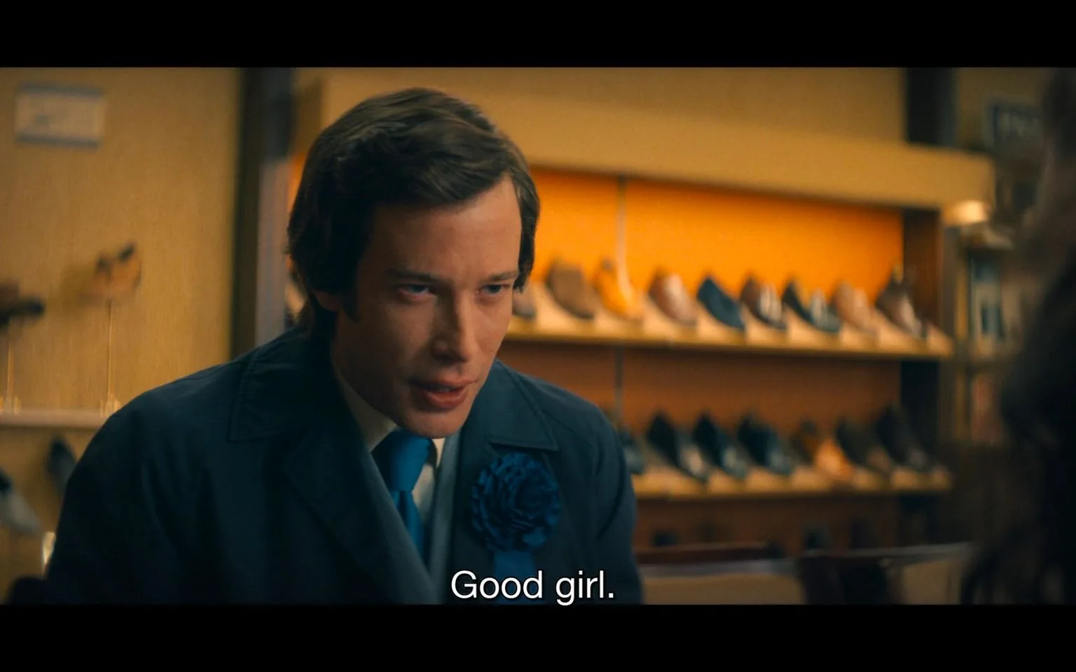 The image captures a scene in a shoe shop, where a man is leaning forward with a focused expression. He is wearing a dark blue suit with a blue tie and a large blue flower pin on his lapel. The background features a display of various shoes, all neatly arranged on shelves with warm lighting creating a cozy atmosphere. The man appears to be in conversation, as indicated by the subtitle at the bottom reading, "Good girl." His posture and expression suggest he is engaged and perhaps a bit intense in the moment. The setting indicates a commercial or retail environment, with the focus on the interaction taking place. The overall mood of the scene is intimate, with a sense of direct engagement between the characters involved.