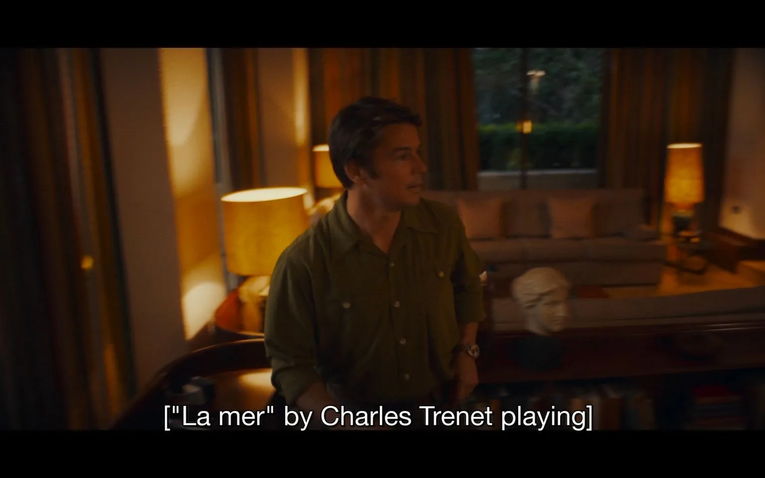 The image depicts an indoor setting, likely a living room, with a warm and cozy atmosphere. A man is seen in the center of the scene, wearing a green button-up shirt. He appears to be in motion, possibly walking or turning, with a neutral expression on his face. The room is softly lit by several lamps, creating a calming ambiance.
In the background, there is a sofa with cushions, positioned beside large windows that offer a view of greenery outside. A sculpture of a bust sits on a table, adding a touch of artistic decor to the room. The room’s design suggests a comfortable and stylish home environment. The lighting highlights the warm earth tones of the room, emphasizing a sense of relaxation.
Captions at the bottom of the image indicate that the song "La mer" by Charles Trenet is playing, contributing to the tranquil mood. The setting and posture of the subject suggest a casual, perhaps introspective moment within a domestic space.
- Captions / subtitles: "["La mer" by Charles Trenet playing]"