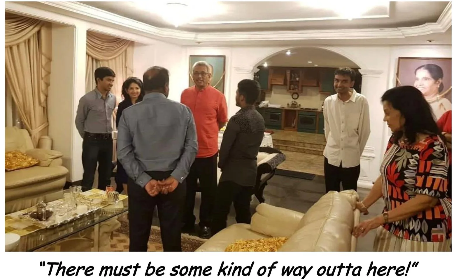 The image depicts a group of people gathered in a well-furnished living room. In the center, Gotabaya Rajapaksa is seen wearing a red shirt, engaging in conversation with others around him. Among the group is Lehan Edirisinghe, who is noticeable due to his direct gaze toward the viewer. The setting is elegant, with light-colored curtains and cushioned furniture, creating a warm and inviting atmosphere. The background reveals a kitchen area, adding to the domestic setting.
The people are casually dressed, suggesting an informal gathering. The mood appears lighthearted and social, with some individuals smiling. A table with glasses and a tray is visible, indicating refreshments might be part of the meeting. The lighting is bright and even, enhancing the room's welcoming ambiance.
Below the image is a caption reading, "There must be some kind of way outta here!"—a line from the song "All Along the Watchtower" by Jimi Hendrix and Bob Dylan, adding a layer of interpretive intrigue to the scene.