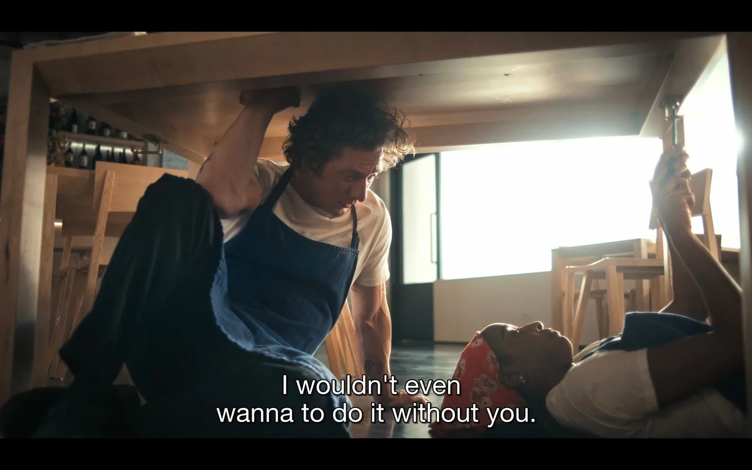 In this scene, two individuals are underneath a wooden table in a restaurant setting. The man is wearing a white t-shirt and a blue apron, suggesting he works in a culinary environment. He is seated with one arm bracing against the table, looking towards the woman next to him. The woman is lying on the floor, holding something in her hand, possibly a phone. She wears a white t-shirt, a blue apron, and a red bandana with white patterns. The lighting is bright, with sunlight streaming through a window in the background, creating a warm, inviting atmosphere. Chairs are stacked neatly in the background, indicating the restaurant may be closed or preparing to open. The subtitle reads, "I wouldn't even wanna do it without you," highlighting a moment of camaraderie or partnership between the two characters.