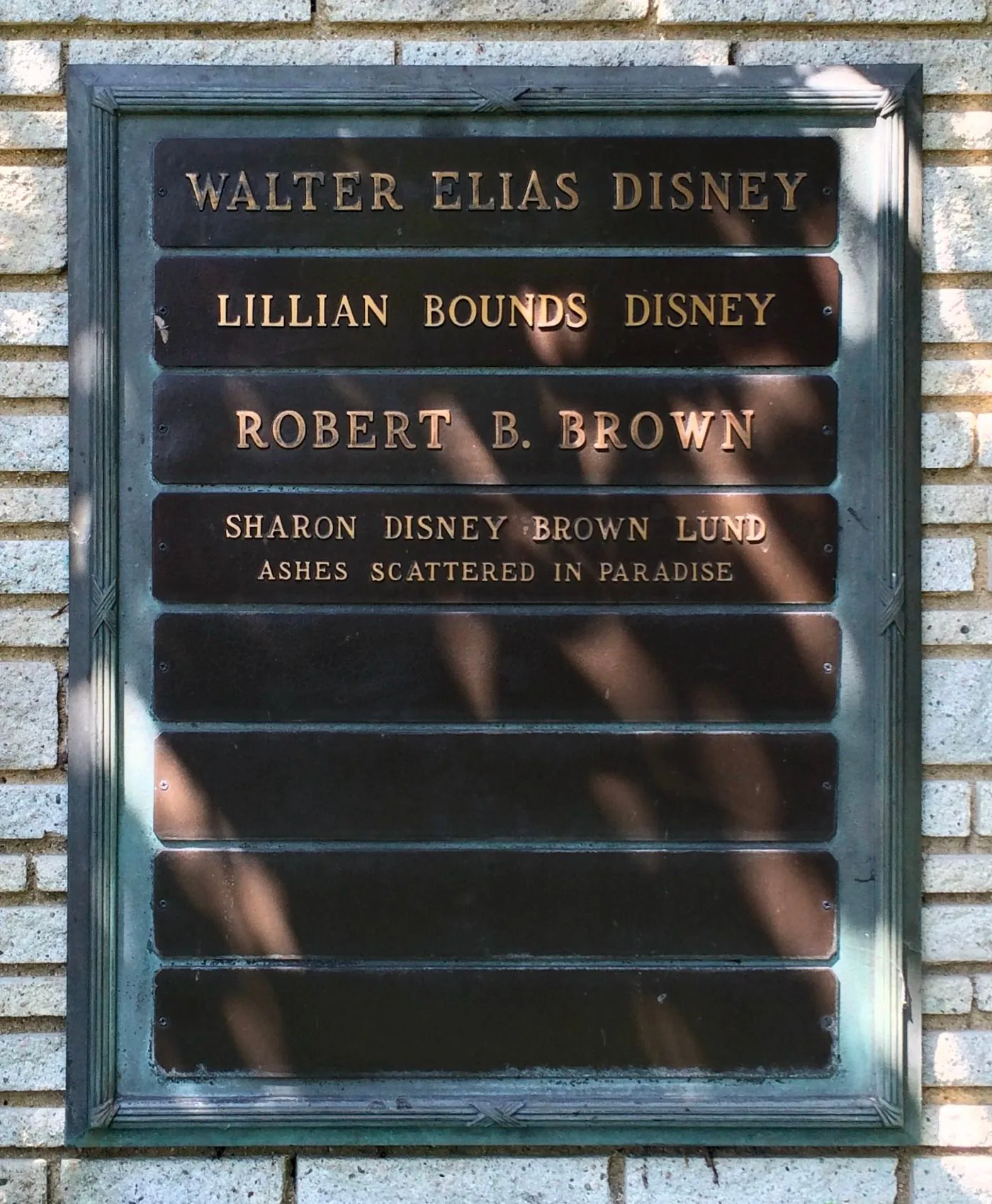 This image shows a memorial plaque set against a brick wall, dedicated to several individuals, including Walt Disney. The plaque is rectangular and made of metal, with names engraved on it in bold, capitalized letters. The names listed include "WALTER ELIAS DISNEY," emphasizing the significance of Walt Disney as the central figure honored here. Below his name, "LILLIAN BOUNDS DISNEY" is also inscribed, indicating a familial connection. Further down, the names "ROBERT B. BROWN" and "SHARON DISNEY BROWN LUND" appear, with a note that her ashes are "SCATTERED IN PARADISE."
The setting is likely outdoors, as indicated by the natural light casting shadows on the plaque. The stone bricks surrounding the plaque add a solemn and respectful tone, fitting for a memorial. The letters are neatly aligned, and the overall presentation is one of dignity and remembrance. The lighting highlights the engraved text, making the names and accompanying notes clearly visible against the darker background of the plaque.