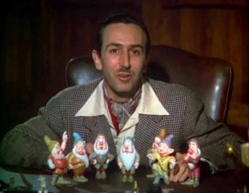 Walt Disney is seated at a desk, appearing younger, in an indoor setting. He is dressed in a checkered suit jacket with a white shirt and a patterned scarf, exuding a mid-20th-century style. In front of him on the desk, there are figurines of the seven dwarfs from the classic animated film "Snow White and the Seven Dwarfs." These figurines are arranged neatly, showcasing characters like Sleepy, Grumpy, Doc, and others, each with distinctive facial expressions and colorful outfits. The background features wooden paneling, adding a warm and cozy atmosphere to the scene. Walt Disney looks directly at the viewer, with a slight smile, embodying a sense of pride and enthusiasm, likely discussing or presenting his work. The lighting is soft, highlighting both Disney and the figurines prominently, giving the image an inviting and nostalgic feel.