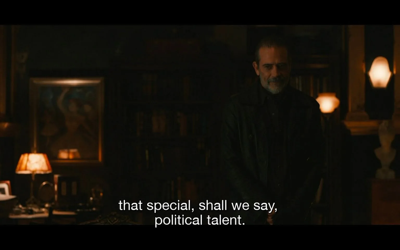Jeffrey Dean Morgan is depicted in this dimly lit indoor scene, wearing a dark leather jacket that adds to the character's rugged appearance. The setting has a moody, dramatic ambiance, emphasized by the subdued lighting and warm tones. Behind him, bookshelves filled with various volumes create a scholarly atmosphere, while a few lamps cast a soft glow, highlighting the room's classic decor. A framed painting is visible on the wall, contributing to the room's sophisticated aesthetic. The scene captures Morgan in a thoughtful pose, with his hands clasped and a contemplative expression, suggesting a moment of introspection or plotting. The subtitle at the bottom of the screen reads, "that special, shall we say, political talent," hinting at a discussion or narrative involving political intrigue or strategy.
- Captions / subtitles: "that special, shall we say, political talent."