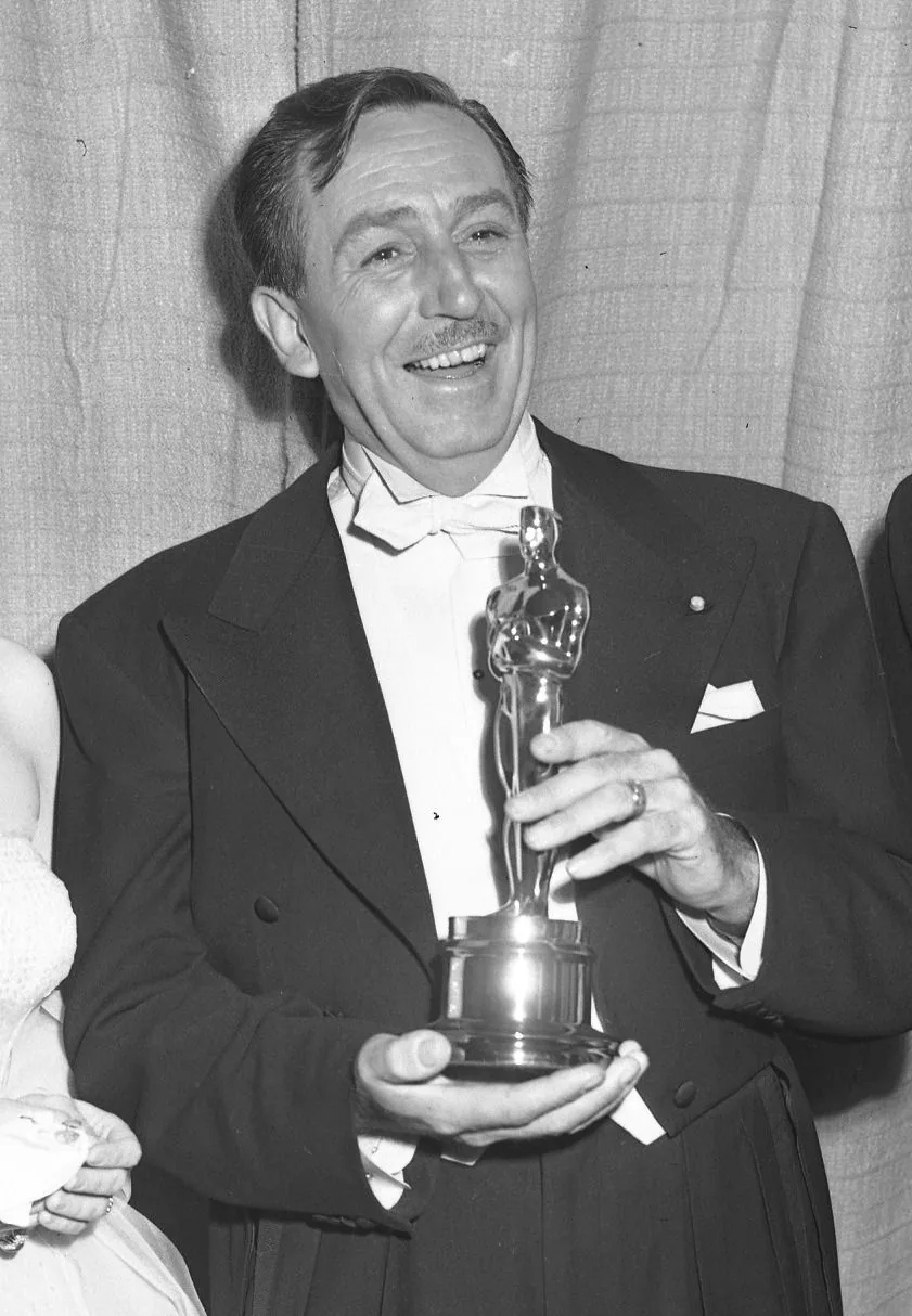 Walt Disney is seen in this black and white photograph, captured in a formal setting, likely an award ceremony. He is dressed in an elegant tuxedo with a white bow tie and pocket square. His expression is joyful and proud as he holds an Oscar statuette prominently in both hands, showcasing a moment of triumph. The background features a simple curtain, highlighting the focus on him and the prestigious award. This image captures a timeless moment of recognition and achievement in the entertainment industry, with Walt Disney exemplifying a sense of accomplishment and pride. The lighting is soft, enhancing the celebratory atmosphere of the scene. The photograph, with its classic and historical feel, conveys both the personal success of Walt Disney and his significant contributions to film and animation.
