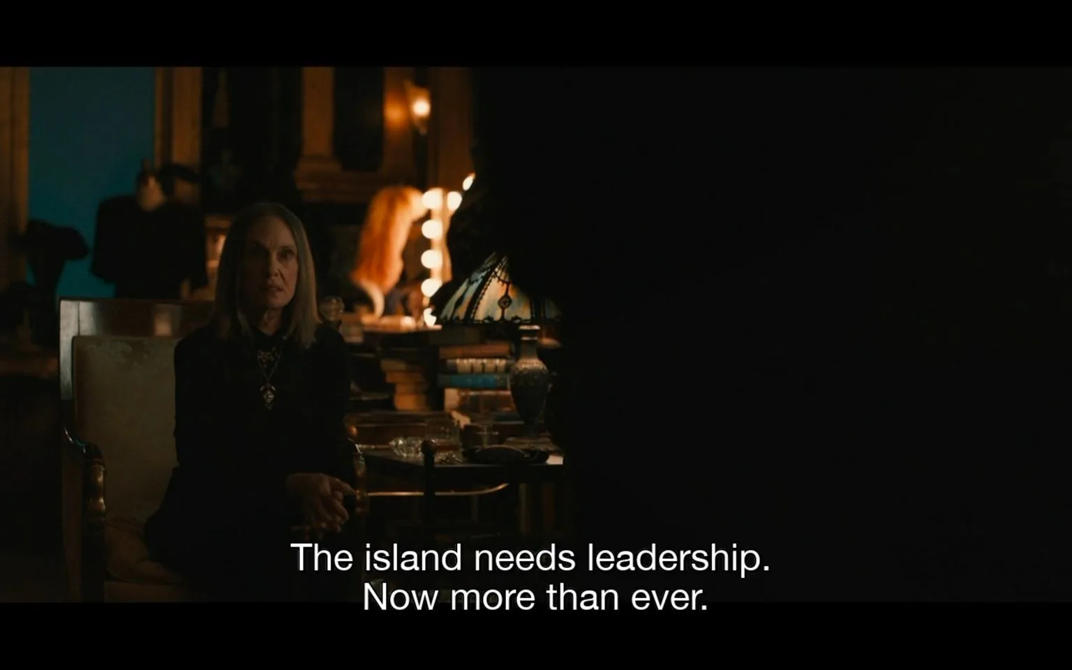 The image is a dimly lit indoor scene featuring a woman sitting in a room that has an ornate and somewhat antique ambiance. She is positioned on the left side of the frame, sitting on a cushioned chair. Her attire is dark, possibly black, which complements the moody atmosphere of the setting. Her posture suggests attentiveness or seriousness. In the background, there's a dressing table with illuminated vanity lights, hinting at a possibly theatrical or vintage setting. Various objects like books and a decorative lamp add to the room's character, suggesting an eclectic or scholarly environment. The lighting is soft and subdued, casting warm tones across the scene, enhancing the intimate and contemplative mood. The subtitle reads, "The island needs leadership. Now more than ever.," indicating a moment of dialogue that suggests urgency or a call to action.