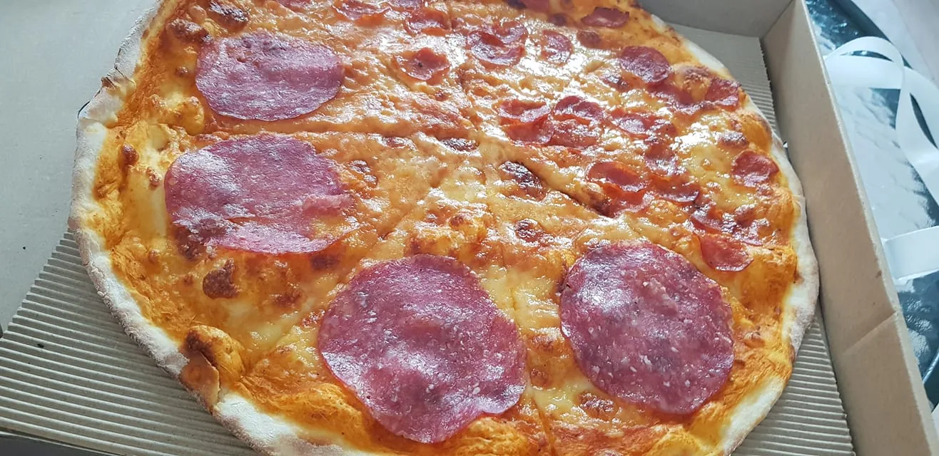 The image shows a pizza in an open cardboard box. The pizza is topped with a layer of melted cheese, and large slices of salami are placed on one half. The crust appears to be thin and slightly crispy, with a light golden-brown color. The cheese is evenly melted across the surface, giving it a rich, appetizing appearance. The cardboard box is standard for takeout pizzas, with ridged edges visible beneath the pizza. The lighting in the image is natural, highlighting the texture and color of the toppings. The setting suggests an indoor, casual dining environment, likely at home or in a casual restaurant setting.