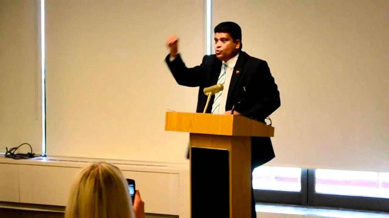 ```
In the image, Shavendra Silva is giving a speech at a podium in an indoor setting. The scene is well-lit, with large windows covered by blinds in the background. Shavendra Silva is wearing a dark suit with a light blue tie, conveying a formal and professional appearance. His expression and hand gesture suggest that he is emphasizing a point during his speech. A microphone is positioned on the podium, indicating he is addressing an audience. In the foreground, an individual with blonde hair is visible, holding up a smartphone, likely recording or taking a photo of the event. The room appears to be a conference or meeting space, with simple and modern decor. The atmosphere is serious and attentive, reflecting the formal nature of the occasion.