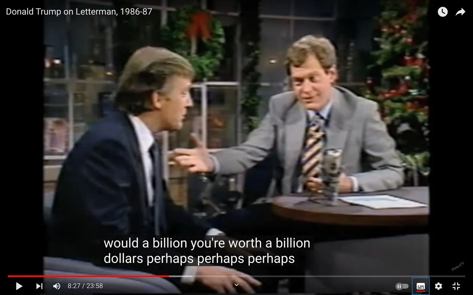 The image is a video frame from a YouTube video titled "Donald Trump on Letterman, 1986-87." It captures a moment from a television interview on the "Letterman" show. The setting is an indoor television studio, featuring a holiday theme with decorations like a Christmas tree and garlands visible in the background.
Donald Trump is seated to the left, wearing a dark suit and tie, engaged in conversation with the host, who is David Letterman. Letterman is on the right side of the frame, dressed in a light gray suit with a striped tie, gesturing with his hand as he speaks to Trump. Both are seated around a small round table, typical of a late-night talk show setup, with a microphone visible on the table.
The lighting is warm and typical of a studio, focused on the interaction between the two men. The mood seems conversational and light, evident from their engaged postures and expressions. There are subtitles at the bottom of the frame, capturing the dialogue: "would a billion you're worth a billion dollars perhaps perhaps perhaps."
- Title: "Donald Trump on Letterman, 1986-87"
- Site / app: "YouTube"
- Captions / subtitles: "would a billion you're worth a billion dollars perhaps perhaps perhaps"