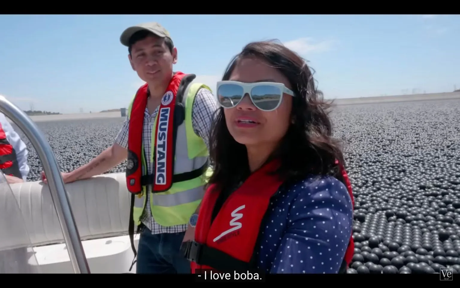 The image shows two people standing on a boat floating in a large body of water covered with numerous black balls, known as shade balls. The scene is outdoors on a sunny day, with clear blue skies above. The woman in the foreground is wearing large reflective sunglasses, a blue polka dot shirt, and a red life jacket. Her expression is engaged and cheerful as she looks towards the camera. The man next to her is dressed in a checkered shirt, a bright yellow safety vest, and a red life jacket with the word "MUSTANG" visible. He is casually resting one hand on the boat's railing and looking in the same direction as the woman. Both are standing amid a vast expanse of these black balls, which are used in reservoirs to reduce evaporation and prevent algae growth. The caption at the bottom humorously reads, "I love boba," drawing a playful comparison between the balls and boba pearls. In the bottom right corner, a small logo with "Ve" is visible, likely representing the video source.