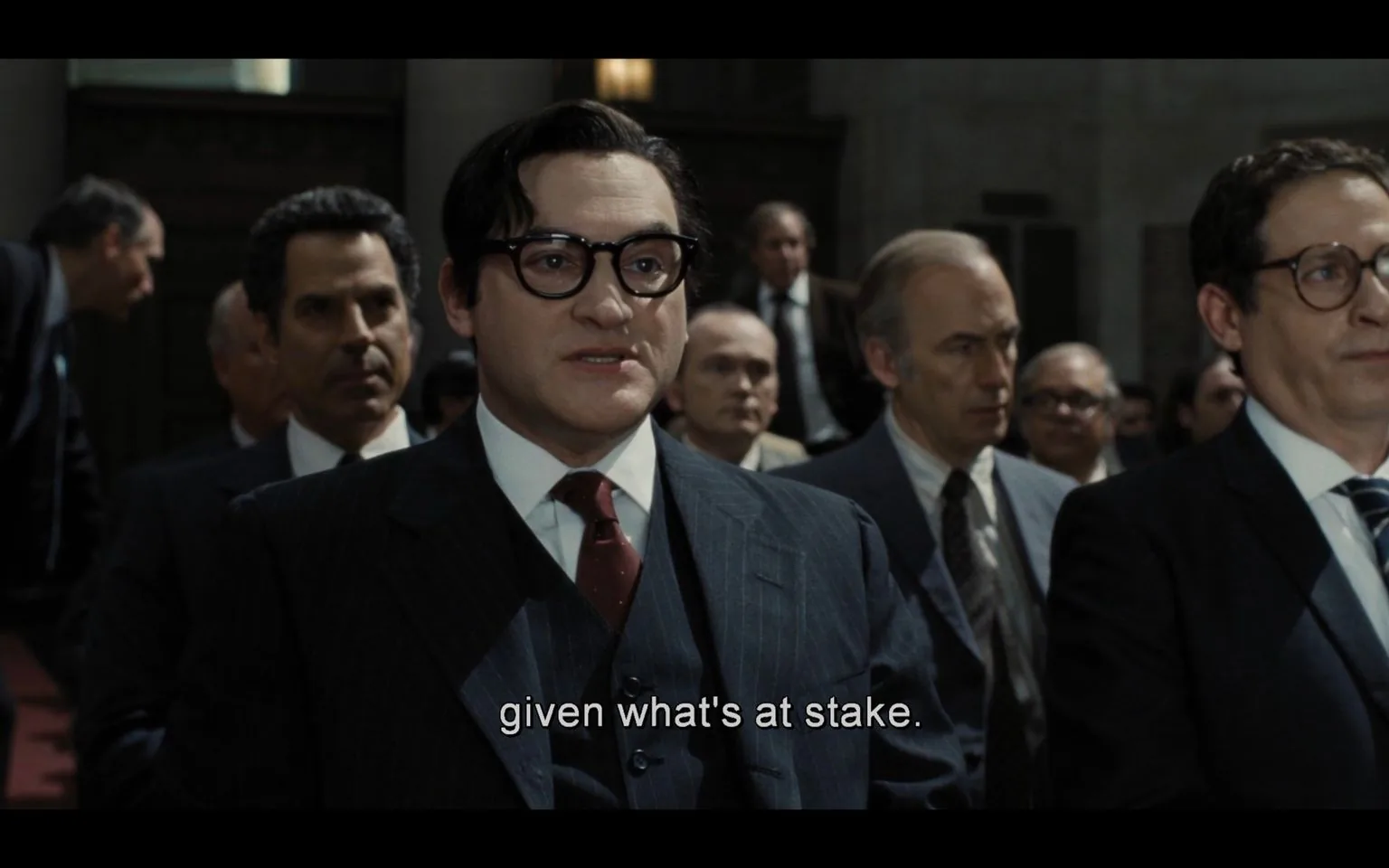 The image is a scene from the film "Oppenheimer," depicting a formal and intense setting within a courtroom or official hearing. The foreground features a central figure, a man in a dark, pinstriped suit and tie, wearing round glasses. His expression appears serious and focused, contributing to the gravity of the moment. Surrounding him are other men, also dressed in formal attire, suggesting a gathering of significant importance, possibly involving legal or governmental proceedings.
The lighting is subdued but clear, emphasizing the formal atmosphere and the tension in the room. The background shows a group of individuals seated closely together, all appearing attentive to the ongoing event. The caption on screen reads, "given what's at stake," hinting at a critical discussion or decision being made. The overall composition suggests a pivotal moment, with the characters deeply engaged in the unfolding events.