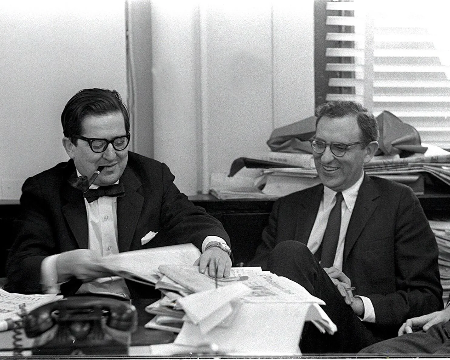 In this black and white photograph, two men are seated in an office setting, engaged in a conversation. On the left, a man with dark hair and glasses, identified as AM Rosenthal, is holding a pipe in his mouth while handling a stack of papers. He is dressed in a formal suit with a bow tie, exuding a professional aura. His expression is focused yet relaxed, suggesting involvement in a thoughtful discussion.
To his right, another man, also wearing glasses and a suit with a tie, is smiling, indicating a lighthearted or agreeable interaction. The table in front of them is cluttered with papers, suggesting a busy work environment, typical of a newsroom or editorial office. A classic rotary telephone is visible in the foreground. The background features a window with blinds partially drawn, contributing to the office atmosphere. The overall mood is one of collaboration and intellectual engagement, characteristic of media and journalism settings of the era.