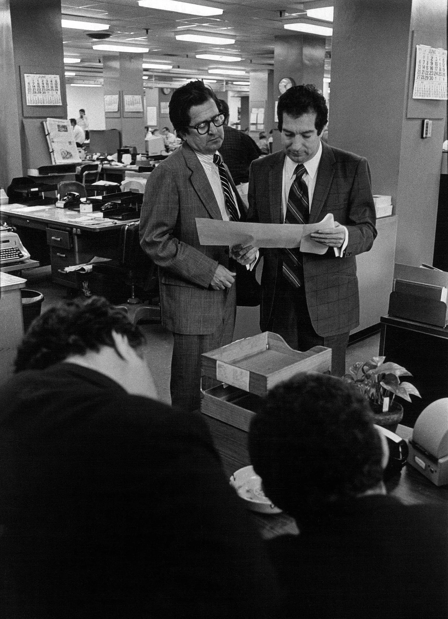 The image depicts an office setting, likely from the mid-20th century, characterized by a black-and-white photograph. The scene captures a bustling workspace with fluorescent lighting and employees engaged in their tasks. In the foreground, two men dressed in suits stand closely together, involved in a discussion. One of them is identified as AM Rosenthal. They are examining a document intently, suggesting a review or decision-making process. Rosenthal wears glasses and a striped tie, while the other man holds a piece of paper and wears a suit with a checkered pattern.
In the background, several desks are visible with typewriters, telephones, and paper stacks, typical of a busy office environment. The decor includes wall calendars, and a clock can be seen, adding to the period-specific details. The atmosphere appears to be serious and focused, with two other individuals slightly out of focus, possibly engaging in their own work at a nearby table. The overall composition conveys a sense of professionalism and the collaborative nature of office work.
