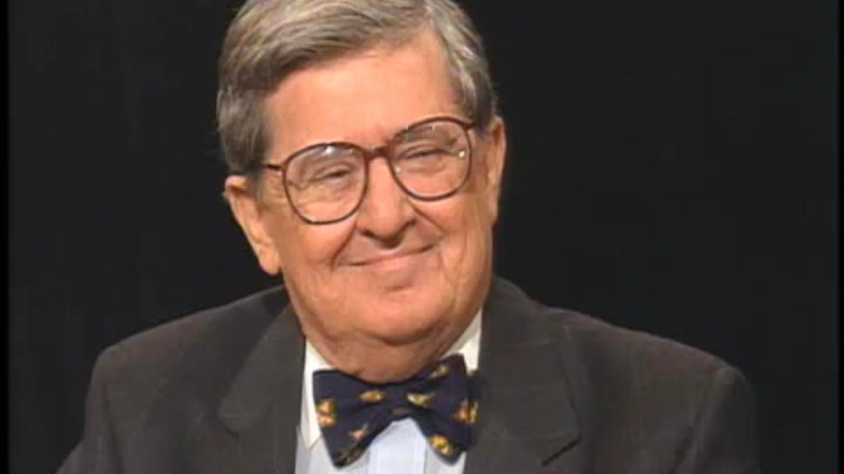The image features AM Rosenthal in a studio setting. He is wearing a dark suit with a light-blue shirt and a patterned bow tie, giving off a formal and polished appearance. His expression is warm and approachable, with a slight smile, suggesting a relaxed and conversational mood typical of an interview or discussion. The background is a plain black, which helps to focus attention on Rosenthal himself. His hair is gray and neatly styled, and he is wearing large, round eyeglasses. The lighting is soft and even, highlighting his facial features without creating harsh shadows. The overall composition is centered, with Rosenthal's head and shoulders prominently displayed, indicating this is likely a still from a television interview or a similar on-camera appearance.