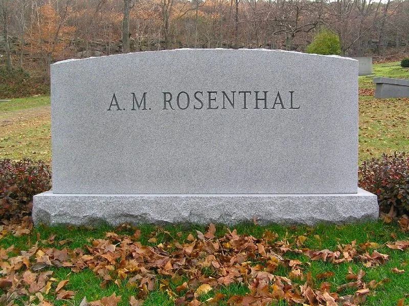 This image depicts a tombstone with the name "A. M. Rosenthal" engraved prominently on its surface. The tombstone is made of a light gray stone and has a simple, slightly arched shape, giving it a traditional appearance. It is set in a well-maintained cemetery surrounded by grass and fallen autumn leaves, suggesting a serene and peaceful environment typical of the fall season. Behind the tombstone, there are trees with bare branches, indicating late autumn or early winter. The background also includes another tombstone, partially visible, contributing to the tranquil and respectful atmosphere of the scene. The overall lighting is subdued, consistent with an overcast day, enhancing the reflective mood of the setting.