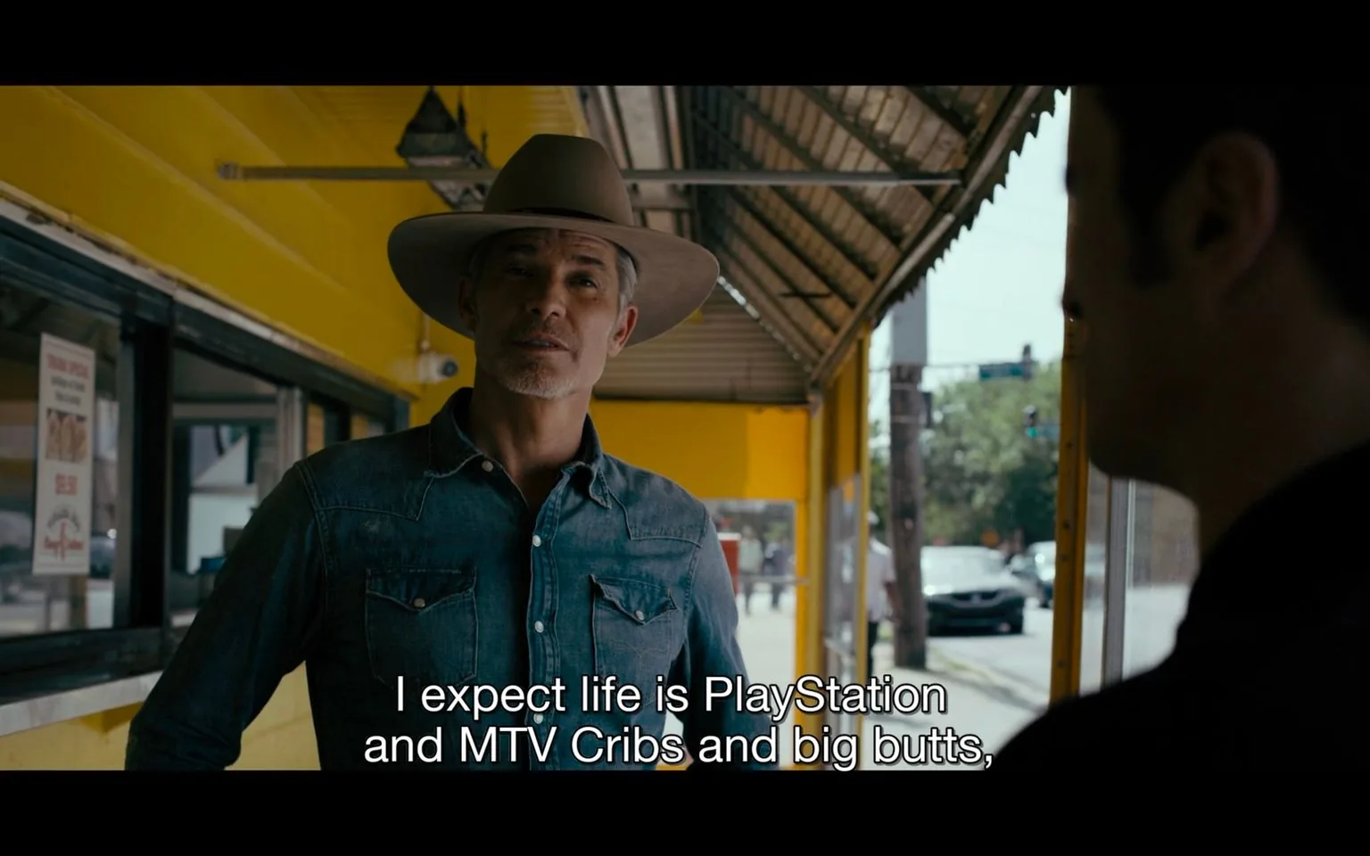 The image depicts an outdoor scene with two individuals engaged in conversation. One person, wearing a wide-brimmed hat and a denim shirt, stands in front of a brightly painted yellow building. His expression appears thoughtful as he addresses the other person. The building has a corrugated metal overhang, casting a shadow over the scene, suggesting a sunny day. A poster with text and pricing is visible on the left, indicating a commercial setting, possibly a diner or small restaurant. The background shows a street with a few vehicles and trees, providing an urban or suburban context. The subtitle at the bottom reads, "I expect life is PlayStation and MTV Cribs and big butts," adding a touch of humor and modern cultural reference to the dialogue. The overall mood is casual, with a focus on everyday life and interactions.
- Captions / subtitles: "I expect life is PlayStation and MTV Cribs and big butts,"