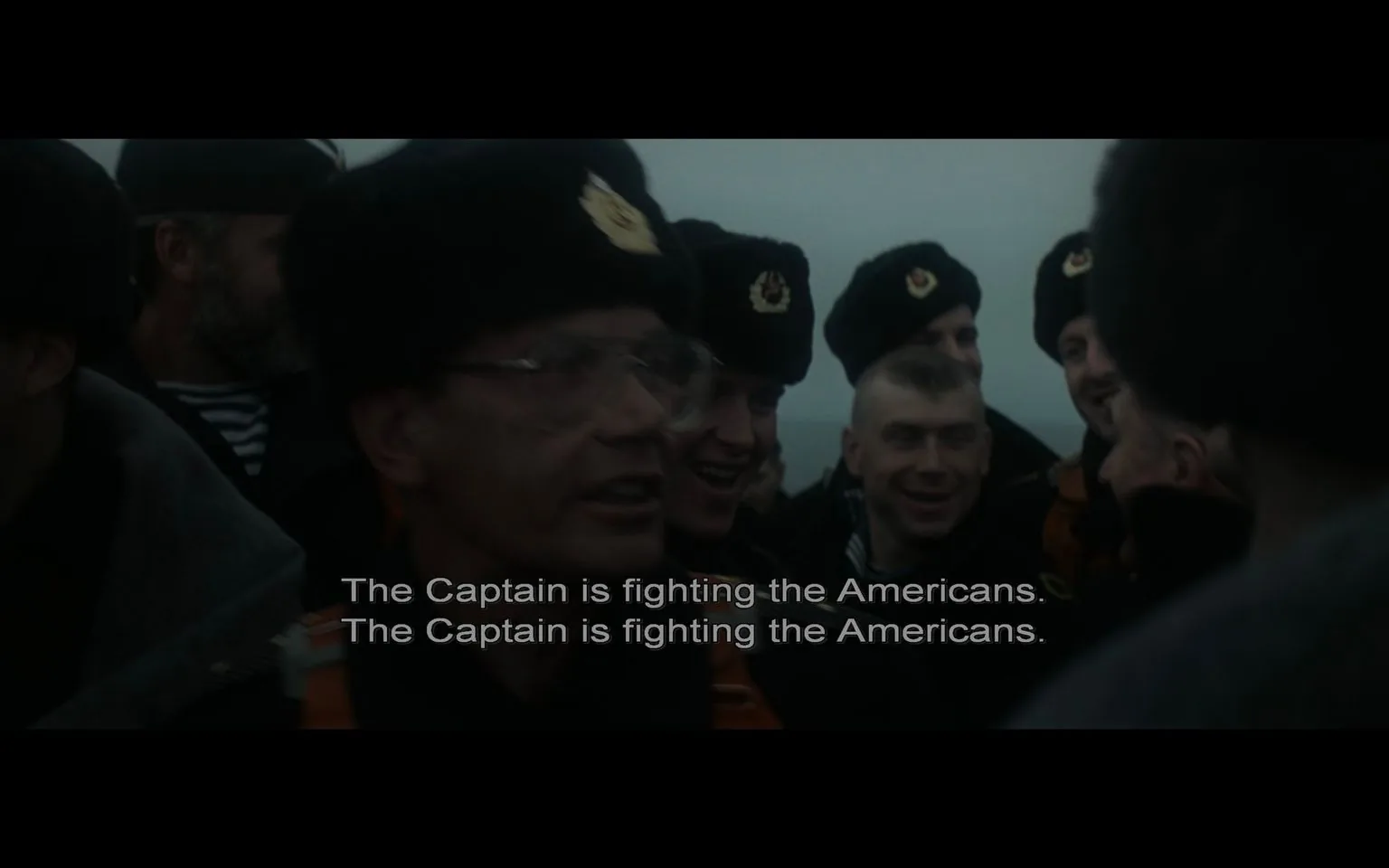 A group of men is gathered outdoors, dressed in military uniforms, indicating a naval setting. They are wearing dark clothing and distinctive fur hats with emblems, suggesting they are part of a Russian naval crew. The expressions on their faces vary from serious to slightly amused, as they appear to be engaged in a conversation or event of significance. The lighting is dim, giving the scene a somber and tense atmosphere, which matches the context provided by the subtitles. The subtitle reads, "The Captain is fighting the Americans," emphasizing a conflict or standoff situation involving a captain and American forces. This aligns with themes from "The Hunt for Red October," a film known for its Cold War naval drama. The image captures a moment filled with tension and camaraderie among the crew members.