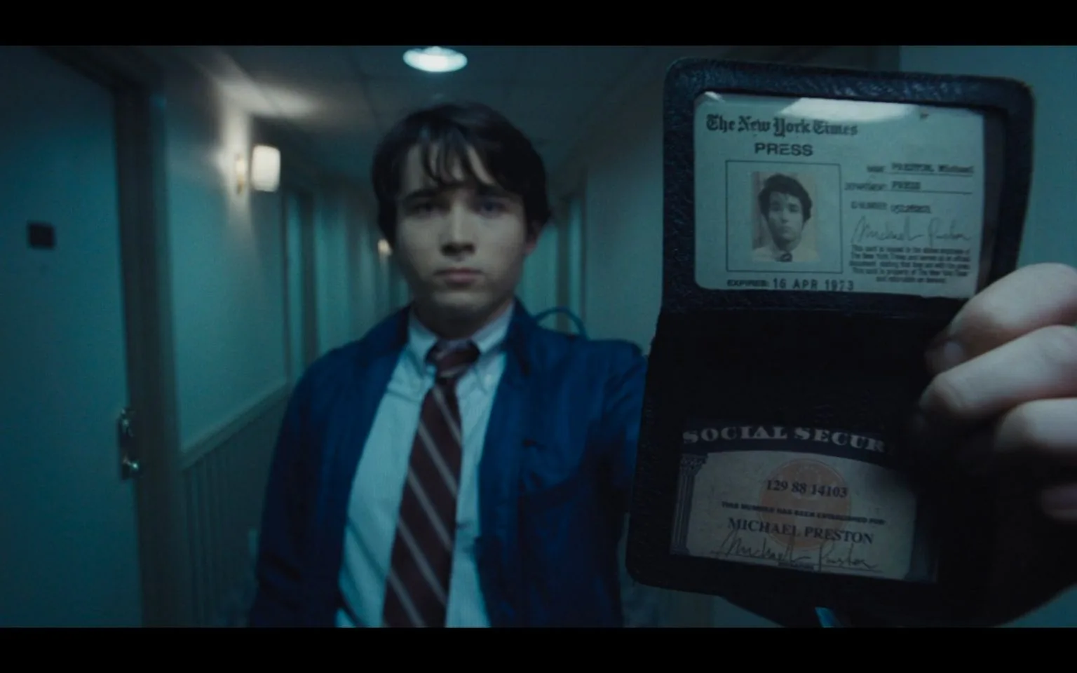 The image depicts an indoor scene in a dimly lit hallway. A young man is in focus, wearing a blue jacket over a white shirt and a red-striped tie. He holds up an identification wallet prominently in the foreground. The ID card inside displays the logo of "The New York Times" and is labeled as a press pass for "Michael Preston," with an expiration date of "16 APR 1973." Below it, a Social Security card is visible, also bearing the name "Michael Preston" and the number "129 88 14103."
The hallway appears narrow, with closed doors along the right side and a few wall-mounted lamps providing minimal lighting. The mood is somewhat tense or serious, possibly due to the setting and the direct way the ID is presented. The focus on the identification suggests a moment of verification or confrontation. The color palette is muted, with blue and gray tones dominating the scene, enhancing the somber, official atmosphere.