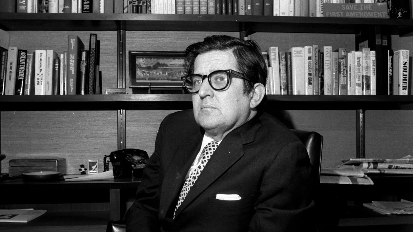 The black and white image shows a man, identified as AM Rosenthal, sitting in an office setting. He is wearing a dark suit, white shirt, and a patterned tie. His expression is serious as he looks slightly off to the side. Rosenthal is seated in front of a bookshelf filled with various books, some of which have visible titles like "Save the First Amendment," "Asian Dream," "The Transition of Israel," and "Soldier." The office has a classic feel with a vintage telephone and neatly stacked papers on the desk. The lighting is subdued, emphasizing the contrast typical of a black and white photograph. The overall mood is professional and contemplative, capturing a moment of introspection or thought.
- Title: "AM Rosenthal in His Office"