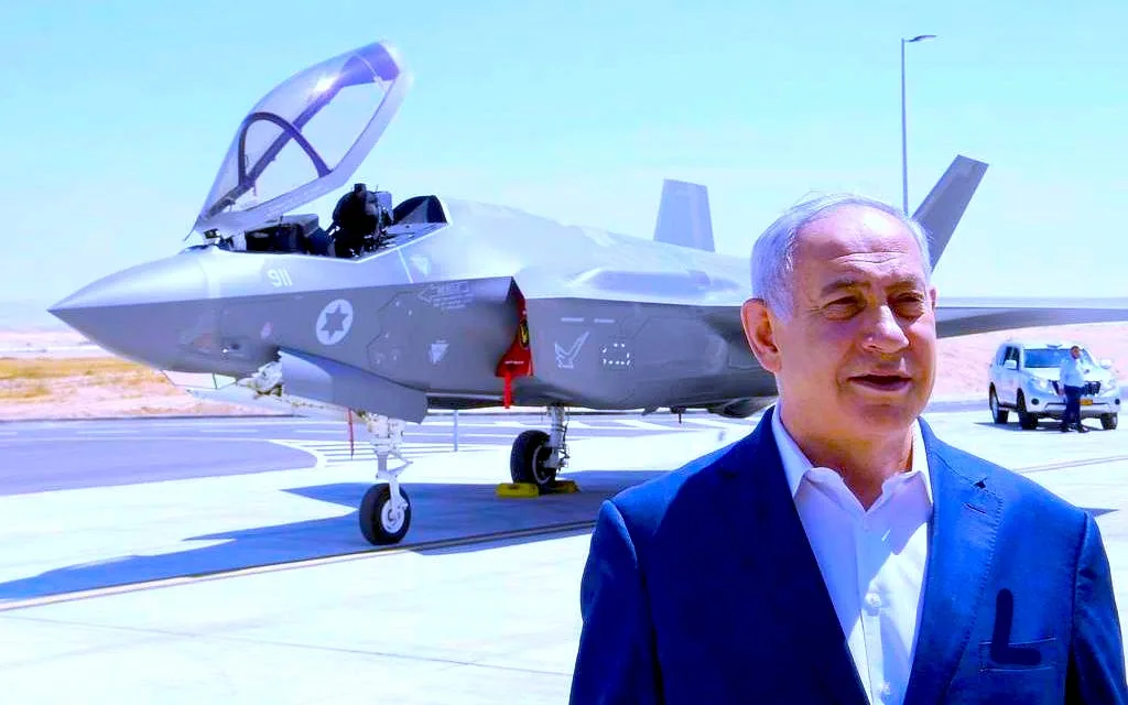 Benyamin Netanyahu is prominently featured standing in front of a modern military jet on a clear, sunny day. The aircraft, an F-35 fighter jet, has its canopy open and displays the insignia of the Israeli Air Force on its side. Netanyahu is wearing a formal suit, with a white shirt underneath, and appears to be at ease, with a slight smile. In the background, a desert-like landscape can be seen, along with a couple of vehicles parked on the tarmac. The lighting is bright, highlighting the sleek, aerodynamic design of the jet. The scene suggests an official or ceremonial context, possibly related to a military event or announcement involving the aircraft.