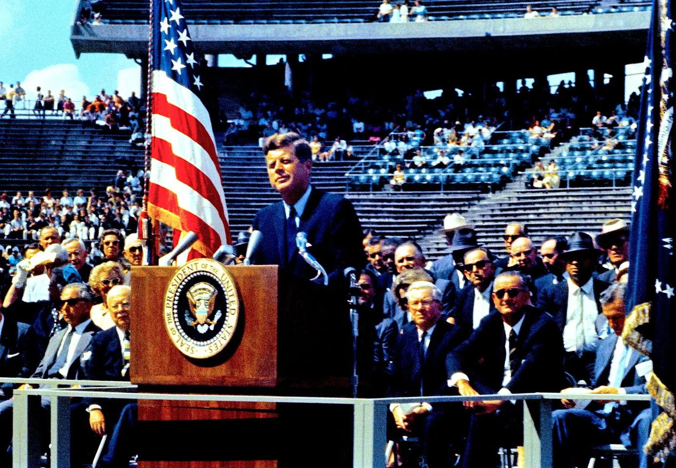 In this historical image, JFK is delivering a speech at a stadium, likely during a significant political or public event. He stands behind a podium adorned with the Seal of the President of the United States, indicating his official capacity. The American flag is prominently displayed beside him, adding a patriotic element to the scene. JFK appears focused and composed, wearing a dark suit and tie, typical of his presidential appearances.
The audience is comprised of men in suits, some wearing sunglasses and hats, suggesting the formal nature of the occasion. The stadium setting is partially filled with spectators, some seated and others standing, likely attentive to the speech. The bright daylight casts clear shadows, enhancing the vivid colors of the event, particularly the red, white, and blue of the flag. This image captures a moment of historical significance, reflecting JFK's role in pivotal national or international discussions, possibly related to the space race or other major initiatives of his presidency.