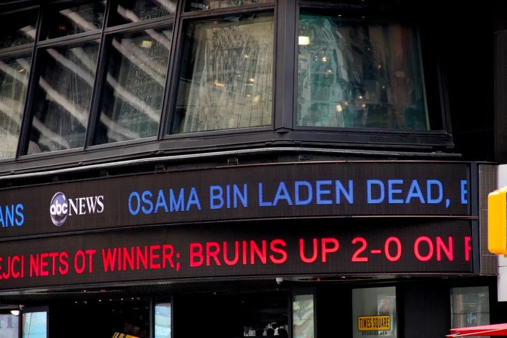The image depicts a news ticker on the exterior of a building displaying breaking news headlines. The prominent text reads "OSAMA BIN LADEN DEAD," indicating a major news announcement about the death of Osama bin Laden. This text is in blue, making it stand out against the black background of the ticker. Below, in red text, a sports update reads, "BRUINS UP 2-0 ON P," which likely pertains to a playoff series involving the Boston Bruins.
The building is identified as part of ABC News, with their logo visible on the ticker. The setting appears to be outdoors, possibly in a bustling urban area, given the architectural style and the presence of large windows reflecting the surrounding environment. The structure suggests a modern media center, likely in a well-known city location, such as Times Square. The lighting is natural, and the image captures part of a street sign or traffic light pole in the bottom right corner, adding to the urban context. The overall mood is serious and urgent, typical of breaking news scenarios. The design and placement of the ticker are intended to attract public attention quickly and convey important information efficiently.