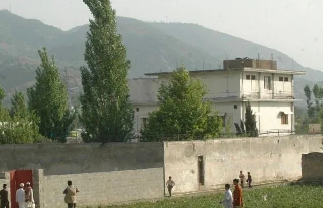 The image depicts a residential area with a prominent two-story building surrounded by a tall boundary wall. The structure appears to be made of concrete, with a flat roof and several windows. It is situated in a lush, green area with trees scattered around, adding to the secluded feel of the location. In the background, rolling hills or mountains are visible, giving a sense of isolation and tranquility. The lighting suggests that the image was taken during the day with natural light illuminating the scene.
In the foreground, a few people can be seen walking or standing near the wall, suggesting casual movement or interaction. Their clothing appears to be traditional, likely indicative of the local culture or climate. The overall atmosphere is quiet and somewhat serene, with no visible signs of urban hustle and bustle. There are no recognizable signs, advertisements, or other textual elements in the image, keeping the focus on the building and its natural surroundings.