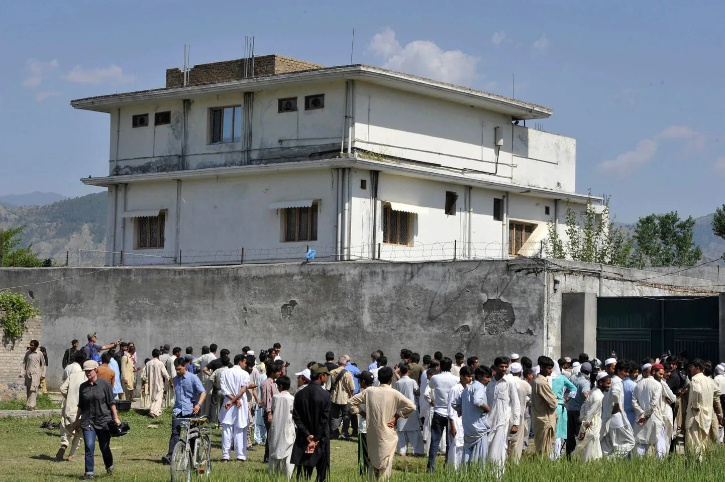This image depicts a large group of people gathered outside a multi-story building surrounded by a tall concrete wall. The building appears to have several barred windows and a flat roof. The crowd is composed of individuals wearing traditional clothing, with a few in more Western attire, suggesting a diverse gathering possibly for an event or a newsworthy occasion. Some people are standing and observing, while others are engaged in conversations, indicating a sense of curiosity or alertness in the atmosphere. The sky is clear with a few clouds, indicating it's a sunny day. The setting appears to be rural or on the outskirts of a town, with mountains visible in the background. The overall mood seems to be one of interest and attention, possibly pointing towards a significant event linked to this location.