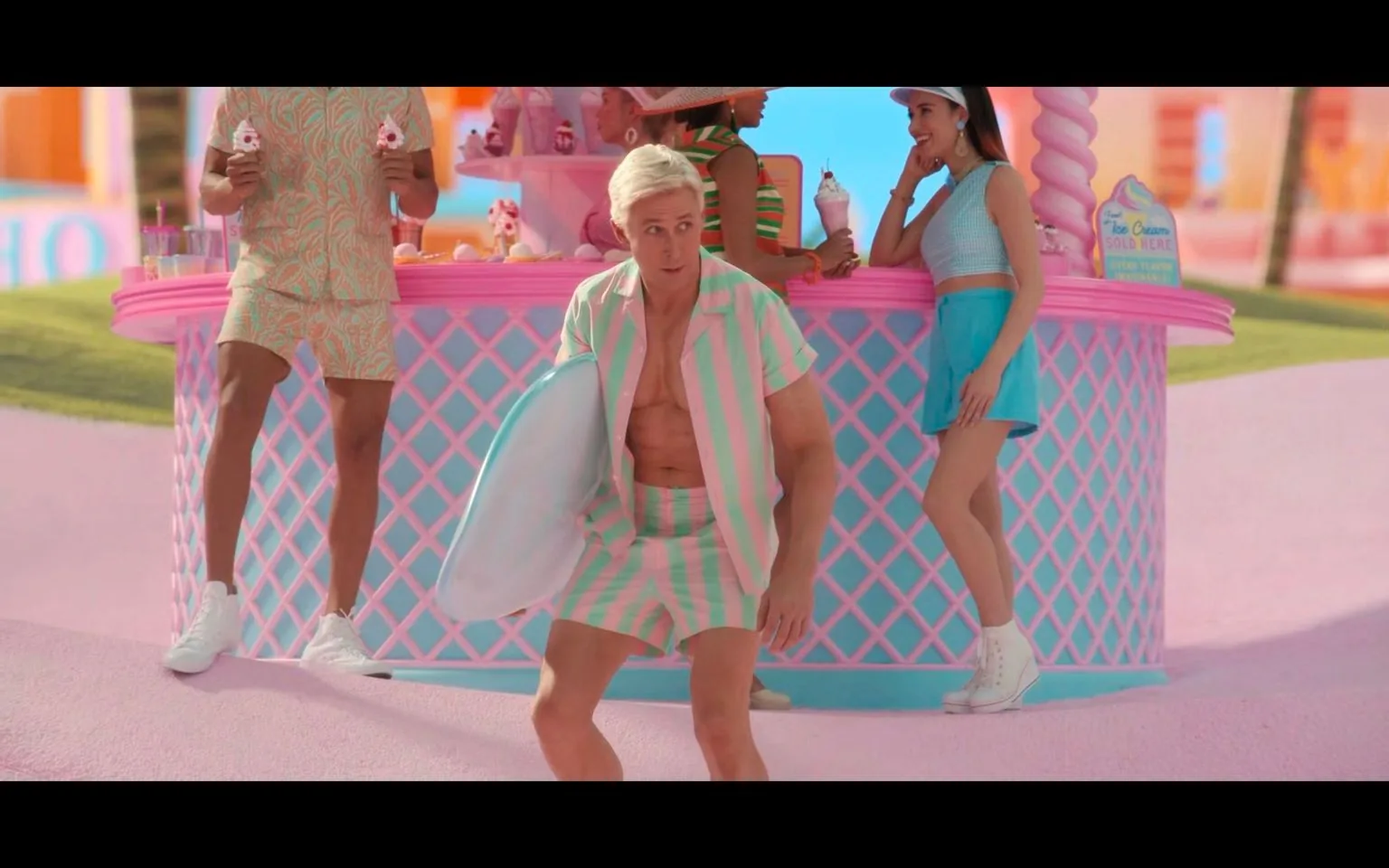The image depicts a vibrant, colorful scene likely from the film "Barbie." In the center, a person resembling Ken is kneeling with a surfboard, wearing a matching striped outfit in pastel pink and green. The background features an eye-catching pink and blue ice cream stand with a checkered pattern. Two other individuals are seen nearby, one holding two ice cream cones and another leaning against the stand, engaging in conversation. The setting is outdoors, with bright, cheerful lighting that enhances the playful, whimsical atmosphere. The overall color palette is dominated by pastel pinks and blues, creating a dreamy, fantasy-like environment. The attire and setting suggest a fun, carefree summer day.