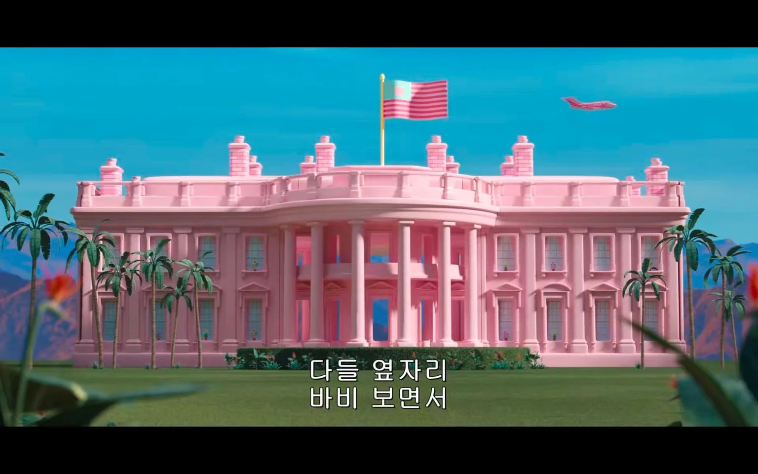 This image shows a large, pink building resembling the White House, surrounded by palm trees. The architecture is classic, with white columns and multiple windows, but the entire structure is painted in shades of pink, creating a playful and whimsical atmosphere. A flag flies at the top, with pink and white stripes, adding to the theme.
The sky is bright blue, and a pink airplane flies in the background. The scene suggests a fantastical setting, possibly in a fictional universe related to Barbie. The landscape is lush with greenery, enhancing the vibrant and cheerful mood. The style is colorful and cartoonish, fitting the aesthetic commonly associated with Barbie.
On-screen Korean text translates to "Everyone looks at Barbie," hinting at the focus on the character in this scene. The overall composition is visually striking, with a blend of realism and stylized elements.