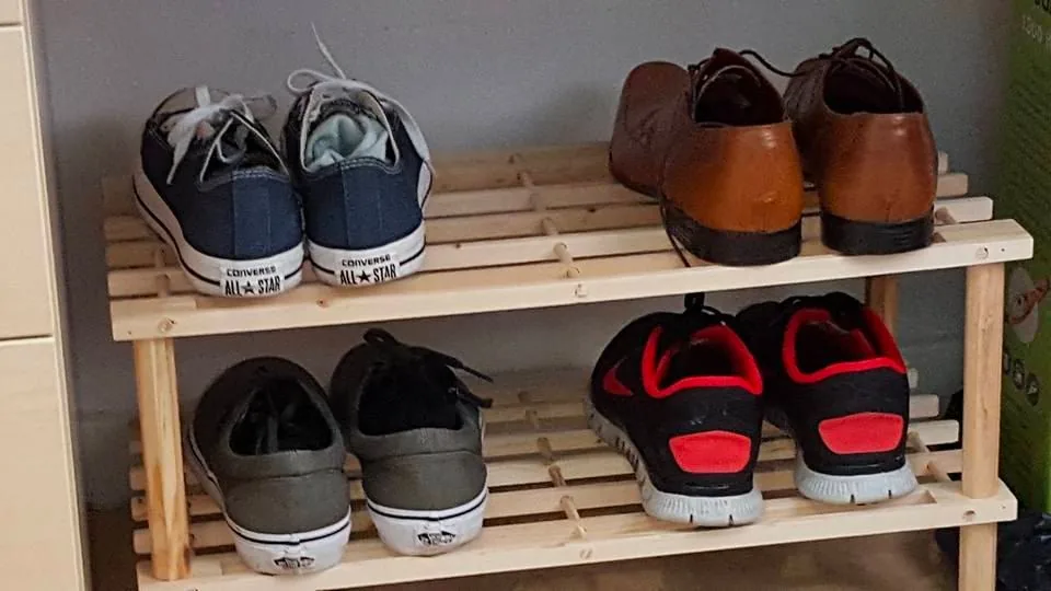 A wooden shoe rack is neatly organized with various pairs of shoes placed on two shelves. On the top shelf, there are two pairs of shoes: a pair of blue Converse All-Star sneakers and a pair of brown leather dress shoes. The Converse sneakers display the iconic branding on the heel. On the lower shelf, there is a pair of gray Converse All-Star sneakers, similar to the ones on the top, and a pair of black and red athletic shoes with a modern design. The shoes are arranged with their heels facing outward, making the brands and styles visible. The setting appears to be a home environment with a simple, functional layout, emphasizing organization and easy access to the footwear. The lighting is soft, casting gentle shadows and highlighting the natural wood color of the rack.