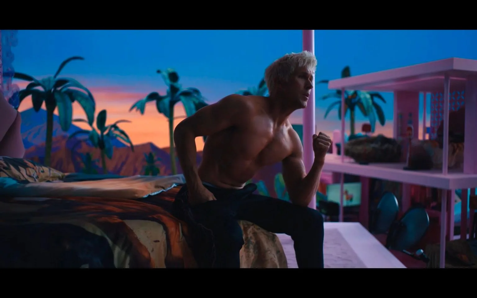 The image features a shirtless man, referred to in the hint as Ryan Gosling, sitting on the edge of a bed. His posture is tense, with one arm slightly bent and fist clenched, suggesting anticipation or determination. The setting is colorful and stylized, likely from a film named "Barbie." The room is vibrant, with a sunset view outside, highlighted by palm trees against a vividly painted sky transitioning from orange to deep blue. The interior decor is playful and modern, with pink accents and a multi-level structure resembling a dollhouse. The lighting creates a dramatic atmosphere, casting shadows and emphasizing the contours of the man's physique. The setting suggests a blend of fantasy and reality, adding to the film's likely whimsical tone.