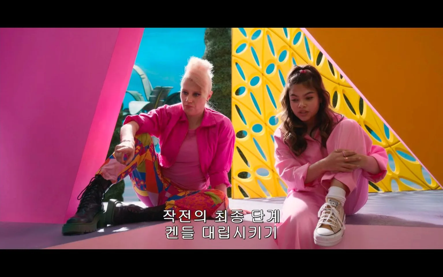 ```
In this vibrant video frame, two individuals are seated in a colorful, geometric environment. The person on the left is wearing a bright pink jacket and multicolored, patterned pants, paired with black combat boots. Their hair is styled in a distinctive, tall mohawk. They appear relaxed, with one arm resting on their knee. To the right, another person is dressed in a pink outfit, including a long-sleeved top and matching pants, accompanied by white sneakers with a visible brand logo. They have long, wavy hair tied back and are hugging their knees. The background is equally colorful, featuring pink and yellow geometric structures with blue accents, creating a playful and dynamic atmosphere. The mood seems casual as if they are in a friendly discussion. The subtitle in Korean reads, "작전의 최종 단계 켄들 대립시키기," which translates to "Final phase of the operation: Incite the Kens against each other."