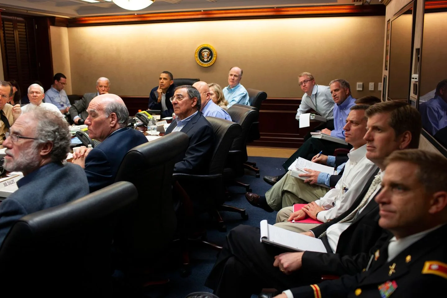 The image depicts a formal meeting taking place in a conference room. The room is filled with several individuals seated around a long, polished wooden table, indicative of a high-level discussion. At the center of the room, a notable seal is mounted on the wall, adding a sense of officialdom to the setting. Most attendees are wearing business attire, such as suits and ties, which suggests a professional context. There are a variety of materials on the table, including notebooks, documents, and telephones, emphasizing the serious nature of the meeting.
The lighting is bright, originating from ceiling fixtures, and the walls are paneled with wood, enhancing the formal atmosphere. Several individuals are taking notes or attentively listening, indicating a focused and engaged environment. A few people appear to be speaking or leading the discussion, while others listen intently. The mood in the room seems serious and concentrated, reflecting the importance of the topics being discussed. The presence of military uniforms suggests that defense or national security might be among the issues on the agenda.