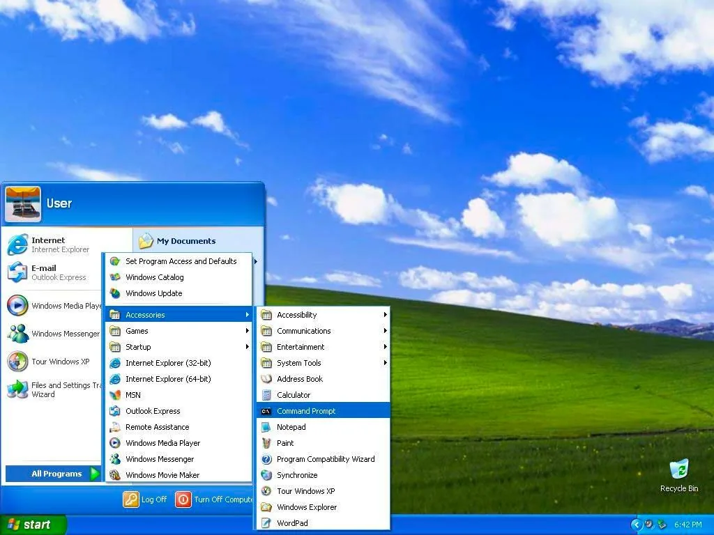The image is a screenshot of a classic Windows XP desktop interface, showcasing a vibrant, iconic background of a green hill under a bright blue sky with scattered white clouds. The central focus is on the Windows XP Start menu, which is open and expanded to reveal several submenus. The Start menu, located at the bottom left of the screen, features a green "Start" button, characteristic of Windows XP.
The menu displays a variety of programs and utilities, including Internet Explorer, Outlook Express, Windows Media Player, and Windows Messenger. Submenu options under "All Programs" include "Accessories," where further selections like "Notepad," "Paint," and "Command Prompt" are visible. The design features a blue theme with white text, typical of the Windows XP user interface. The time displayed at the bottom right is 6:42 PM. The desktop also has a "Recycle Bin" icon, and some system tray icons are visible, indicating system functions and connectivity. The overall look is clean and user-friendly, representing a nostalgic view of early 2000s computing.