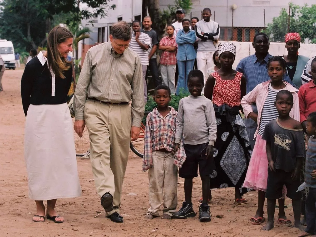 Melinda Gates and Bill Gates are seen in this outdoor scene, walking on a dirt path surrounded by a group of children and adults. Melinda is wearing a black long-sleeve top and a long white skirt, paired with sandals, and she appears to be smiling warmly. Bill is dressed in a light-colored striped shirt and khaki trousers, with black shoes. He is walking with his hands behind his back and also smiling, engaged with the children around them.
The children, dressed in casual clothing, stand in various poses, some looking directly at the camera while others focus on the couple. The background features more people, some with crossed arms, observing the interaction. The setting appears to be in a community area, possibly in a village or small town, with trees and simple buildings visible. The mood is positive and engaging, highlighting a moment of connection and community involvement. The lighting is natural, indicating daytime, and the overall atmosphere suggests a focus on philanthropy and goodwill.