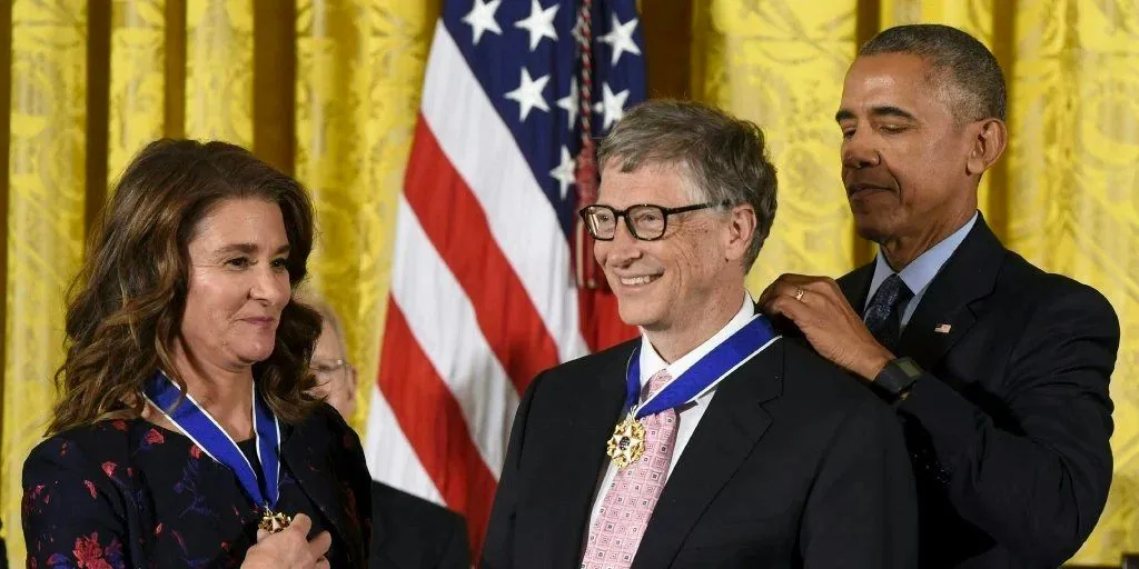 The image depicts a formal awards ceremony set indoors, where Melinda and Bill Gates are seen receiving a medal from President Obama. The setting is elegant, with a richly colored curtain in the background and an American flag displayed prominently. President Obama, positioned on the right, is in the process of placing the medal around Bill Gates' neck. Bill Gates stands centrally, smiling, wearing glasses, a dark suit, and a pink tie. Melinda, standing to the left, is also smiling and wearing a dark dress with a floral pattern. The atmosphere is celebratory and respectful, indicative of the significance of the event. The lighting is bright, highlighting the individuals and their expressions clearly. The medals appear to be the Presidential Medal of Freedom, given their distinctive design.