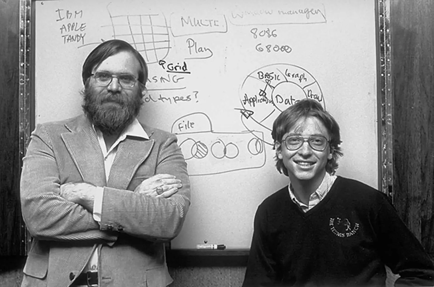 In this historical black-and-white photograph, two individuals are posed in front of a whiteboard filled with diagrams and text related to early computing concepts. The man on the left is standing with his arms crossed, wearing a textured suit jacket and a collared shirt. His expression is serious and contemplative. To the right stands Bill Gates, as indicated by the hint, smiling broadly. He is wearing a dark sweater with writing on it and a collared shirt underneath. Both individuals are wearing glasses.
The whiteboard behind them is cluttered with various technical notes and drawings. Prominent text includes "IBM APPLE TANDY" and terms like "MULTI," "Grid," and "File." There are also references to "8086" and "68000," which are microprocessor models, as well as terms like "Basic," "Application," and "Data," highlighting the focus on early personal computing and software development. The atmosphere is one of innovation and collaboration, capturing a moment in the history of technology development.