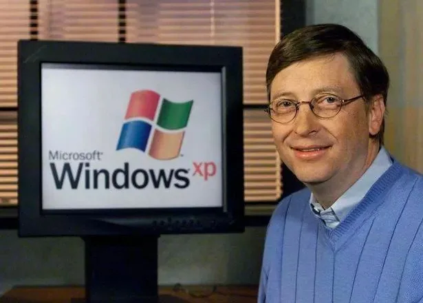 Bill Gates is pictured in an office setting, standing next to a computer monitor displaying the Microsoft Windows XP logo. The logo features the iconic Windows flag in vibrant colors along with the text "Microsoft Windows xp". Bill Gates is wearing a light blue sweater over a collared shirt, and he is smiling pleasantly at the camera. The room has a corporate atmosphere, with blinds partially opened allowing light to softly illuminate the space. The computer monitor and desk suggest a typical early 2000s office environment. The image likely represents a promotional moment for the launch or announcement of Windows XP, emphasizing Microsoft's technological advancements during that era. Bill Gates' relaxed posture and friendly expression contribute to an approachable and professional presentation.