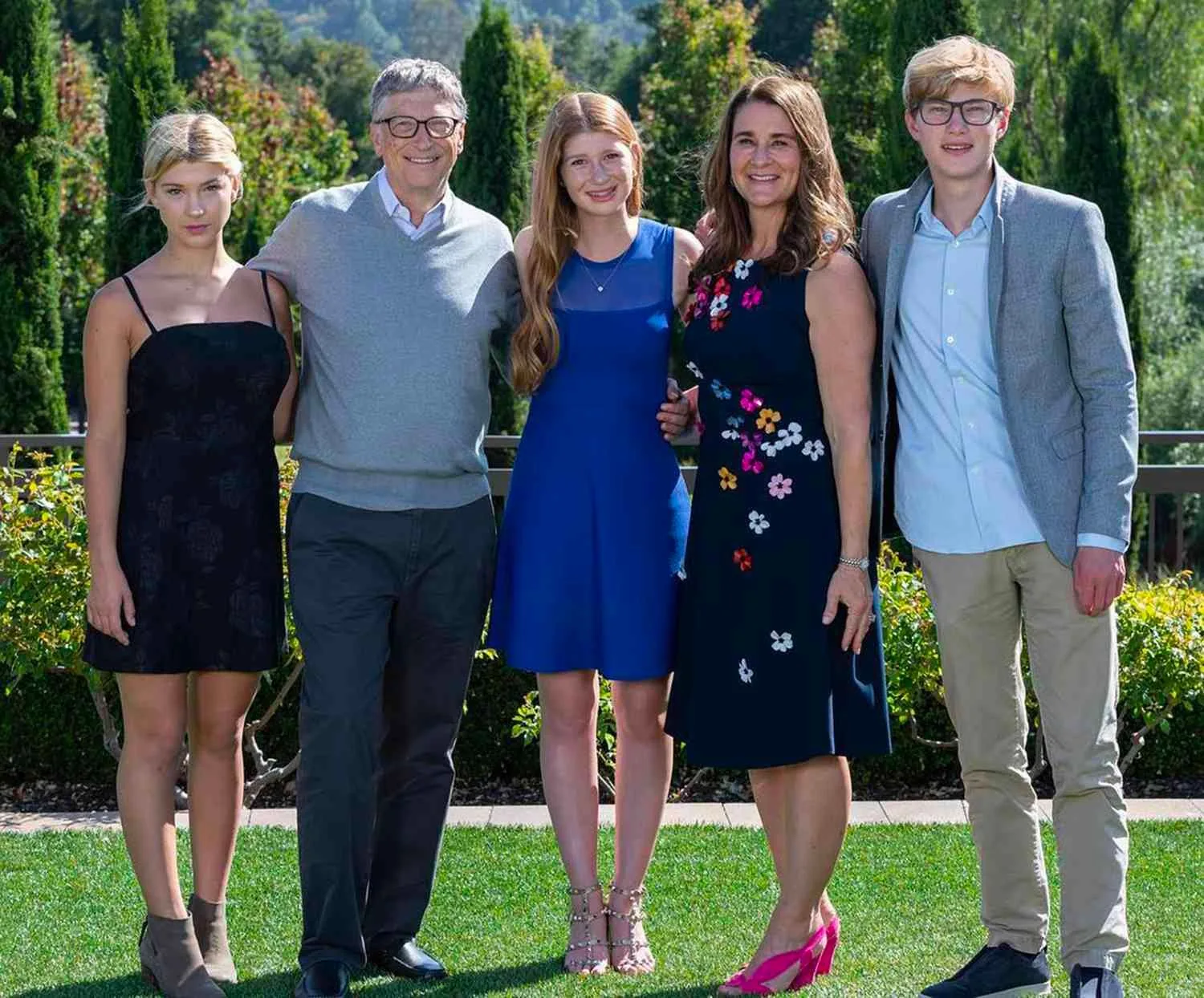 The image features Melinda Gates and Bill Gates with their children, posing outdoors in what appears to be a garden setting. The background is lush with greenery, comprising tall trees and neatly trimmed bushes, which enhances the natural ambiance. The family stands closely together, exuding a warm, united vibe.
Bill Gates, positioned second from the left, is wearing a grey sweater over a white collared shirt, paired with dark trousers. He has a relaxed, approachable demeanor with a slight smile. Melinda Gates, second from the right, is wearing a navy dress adorned with colorful floral patterns and pink heels, adding vibrancy to the scene. Her smile is warm and welcoming, enhancing the familial harmony.
Their children flank them on both sides. The young woman on the left is dressed in a simple black dress and ankle boots, standing with a composed posture. The daughter in the center is wearing a bright blue dress with studded heels, her expression serene and pleasant. The son on the far right is in a light blue shirt, grey blazer, and beige pants, completing the family ensemble with a casual yet neat appearance. The lighting is bright and natural, suggesting a sunny day, which adds a cheerful tone to the overall image.
