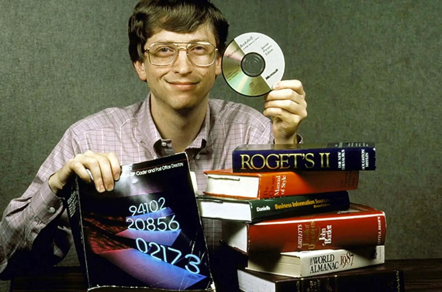 Bill Gates is featured in this vintage portrait, exuding an aura of enthusiasm and curiosity. He is dressed in a light pink plaid shirt, sitting against a plain backdrop. Gates, known for his role in technology, is holding a CD labeled "Microsoft Bookshelf" in one hand, showcasing an early digital product. His other hand rests on a stack of books, including notable titles like "Roget's II," "The New American Webster," and "The World Almanac 1987," illustrating the transition from traditional to digital reference materials.
Gates has a confident smile, suggesting pride in the technological advancements represented by the CD. The lighting is even, highlighting the text on the books and the sheen of the CD. The composition, with Gates centrally placed among the books and media, emphasizes the blend of knowledge and innovation. This image captures a moment in the evolution of personal computing and digital information accessibility.