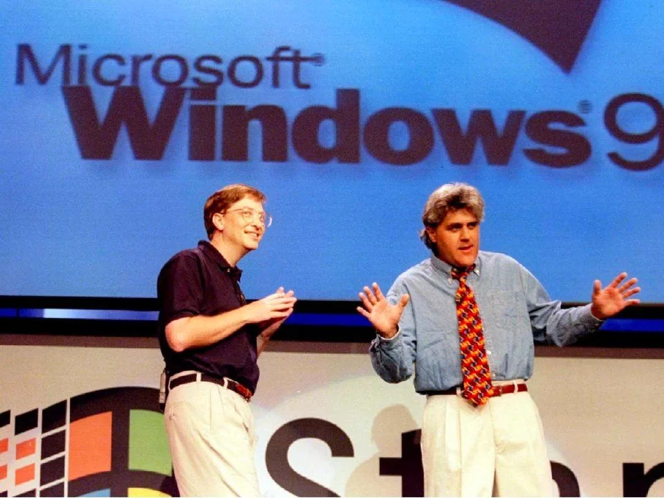 Bill Gates and Jay Leno are on stage during the launch event for Microsoft Windows 95. The backdrop prominently displays the "Microsoft Windows 95" logo, emphasizing the event's focus. Bill Gates is on the left, dressed in a casual, dark polo shirt and light-colored pants, projecting an approachable and relaxed demeanor. Jay Leno stands to the right, wearing a denim shirt and a colorful tie, adding a touch of humor and entertainment to the presentation. Both appear engaged; Gates is smiling and gesturing as he speaks, while Leno has his hands raised in a lively manner, possibly joking with the audience. The stage is brightly lit, highlighting the significance of the technological milestone being celebrated. This image captures a historic moment in technology, blending the technical prowess of Gates with Leno's comedic flair to mark the occasion.